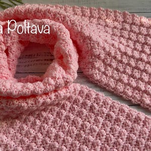 以下が含まれることがあります： 柔らかな、薄いピンク色のかぎ針編みのスカーフ。テクスチャパターンがあります。スカーフは、テクスチャとデザインを示すためにループされています。「(c) Olga Poltava」の文字が左上に表示されています。スカーフは明るい木材の表面にあります。