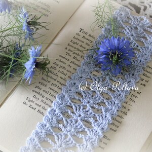 Op de afbeelding: Een blauwe gehaakte kant-bladwijzer met een delicaat bloemenpatroon, liggend op een open boek met blauwe bloemen. De tekst in het boek luidt "THE Blue the Bowery between the t Light does bric-a-brae, it for pain-ki The Blue modern pharm colates its on day pills are r pills rolled ou patula, rolle calcined magu board pill-bos which coveys play and beco soothing syrup Ikey Schoen Light and the East Side, "