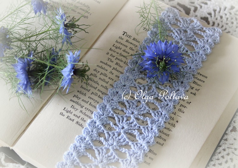 Op de afbeelding: Een blauwe gehaakte kant-bladwijzer met een delicaat bloemenpatroon, liggend op een open boek met blauwe bloemen. De tekst in het boek luidt "THE Blue the Bowery between the t Light does bric-a-brae, it for pain-ki The Blue modern pharm colates its on day pills are r pills rolled ou patula, rolle calcined magu board pill-bos which coveys play and beco soothing syrup Ikey Schoen Light and the East Side, "