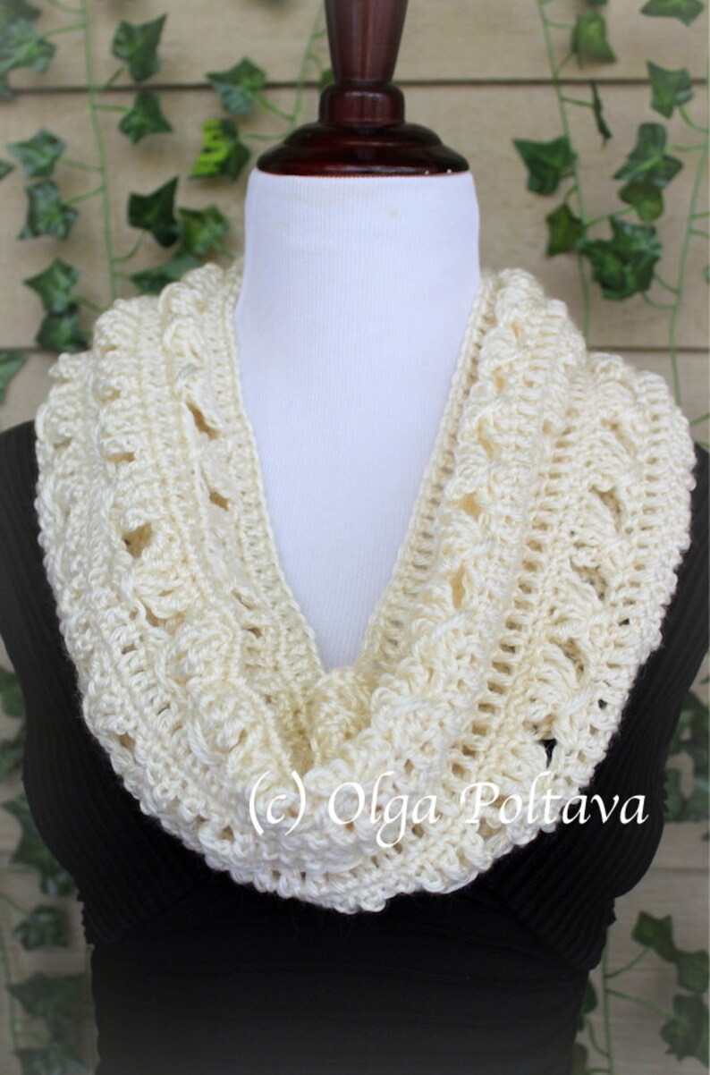 May include: A cream-colored crochet infinity scarf featuring a textured, repeating pattern of raised stitches and openwork loops. The scarf is draped around a white mannequin neck form. The text (c) Olga Poltava is visible on the lower section.