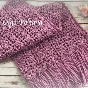 Op de afbeelding: Een gehaakte sjaal in een stoffige roze kleur met franjes aan de onderkant. De sjaal is dubbelgevouwen en ligt op een wit houten oppervlak.