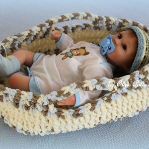 May include: A crocheted baby basket in cream, brown, and blue with a baby doll inside wearing a white onesie with a brown bear design, a blue hat, and a blue pacifier.