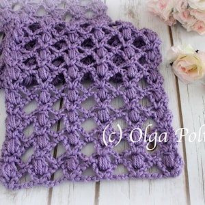 May include: A lilac-coloured, handmade crochet scarf with a textured, openwork pattern. The scarf is folded and displayed on a light wooden surface, with pink roses in the background. The copyright text "(c) Olga Poltava" is visible.