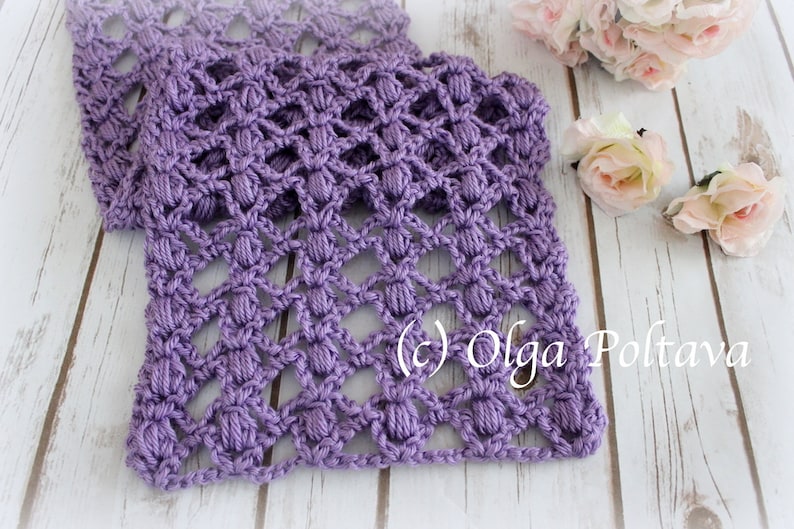 May include: A lilac-coloured, handmade crochet scarf with a textured, openwork pattern. The scarf is folded and displayed on a light wooden surface, with pink roses in the background. The copyright text "(c) Olga Poltava" is visible.