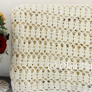May include: A white crocheted throw blanket with a delicate openwork pattern. The blanket is folded and displayed on a table with a vase of flowers.