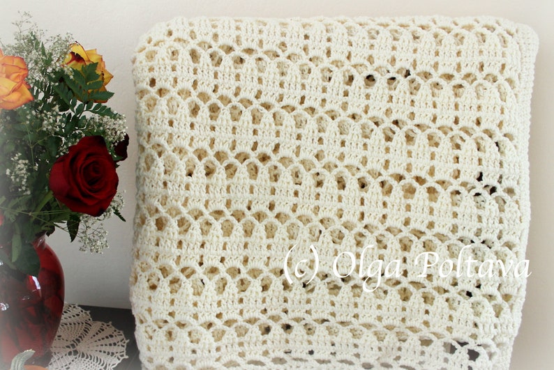 May include: A white crocheted throw blanket with a delicate openwork pattern. The blanket is folded and displayed on a table with a vase of flowers.
