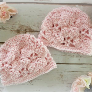 May include: Two light pink crocheted hats with a floral pattern are displayed on a light wood surface. The hats are made of soft yarn and have a delicate, openwork design. Pink roses are in the background, adding a touch of elegance.