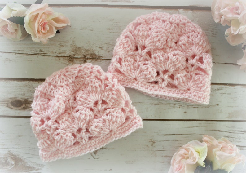 May include: Two light pink crocheted hats with a floral pattern are displayed on a light wood surface. The hats are made of soft yarn and have a delicate, openwork design. Pink roses are in the background, adding a touch of elegance.