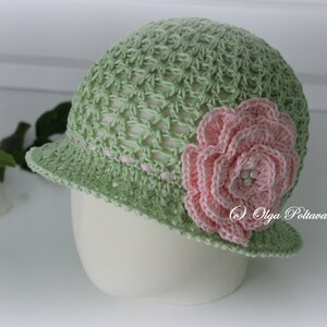 May include: A light green crochet hat with a small brim and a pink flower on the side. The hat has a delicate open weave design and a pink ribbon detail around the brim. The flower has multiple layers and pearl accents.