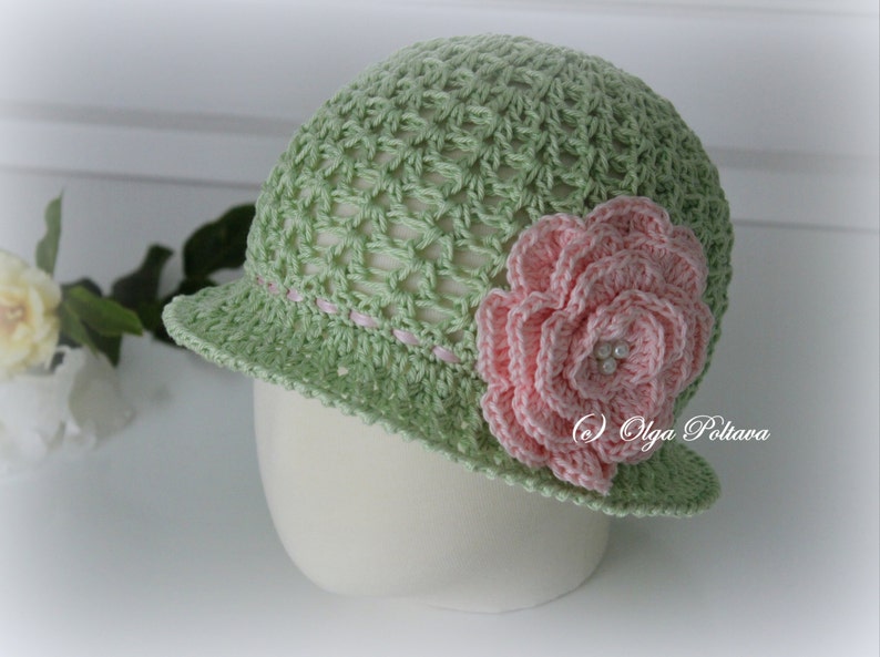 May include: A light green crochet hat with a small brim and a pink flower on the side. The hat has a delicate open weave design and a pink ribbon detail around the brim. The flower has multiple layers and pearl accents.