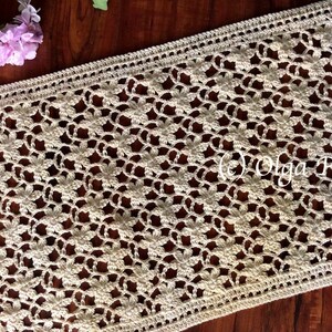 May include: A crocheted table runner with a delicate floral pattern in a cream color. The runner is made with a fine yarn and has a delicate lace border.