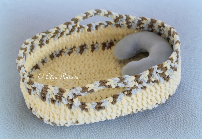 May include: A crocheted basket with brown, blue, and white yarn. The basket has two handles and a light blue pillow inside.