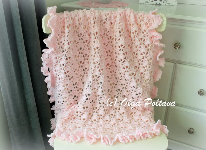 May include: A pink crocheted baby blanket with a delicate lace pattern and ruffled edges. The blanket is draped over a white chair.