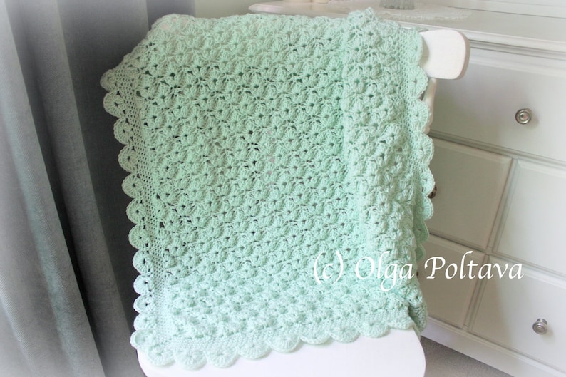 May include: A mint green crocheted blanket with a scalloped edge is draped over a white chair. The blanket features a textured pattern and is made of soft yarn. The blanket is a handmade item, perfect for a baby or as a decorative piece.