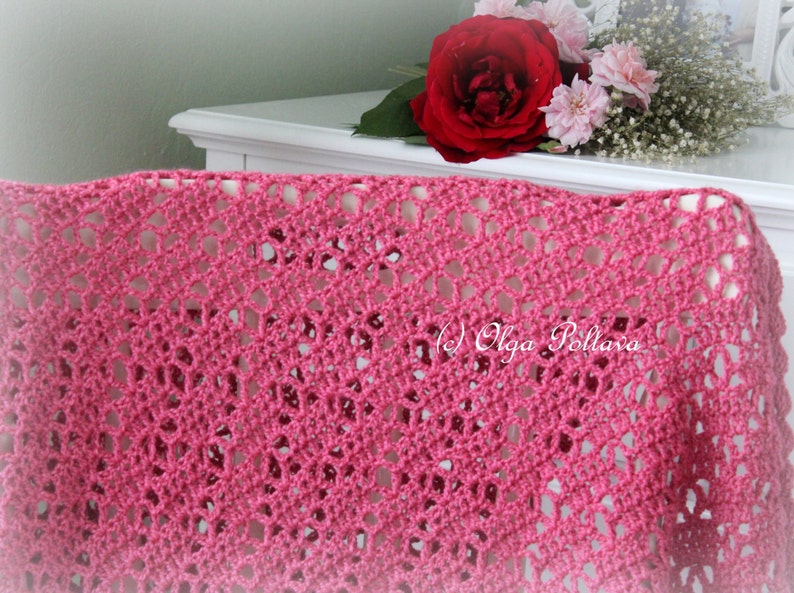 May include: A pink crocheted shawl with a delicate openwork pattern. The shawl is draped over a white surface with a bouquet of red and white roses in the background.