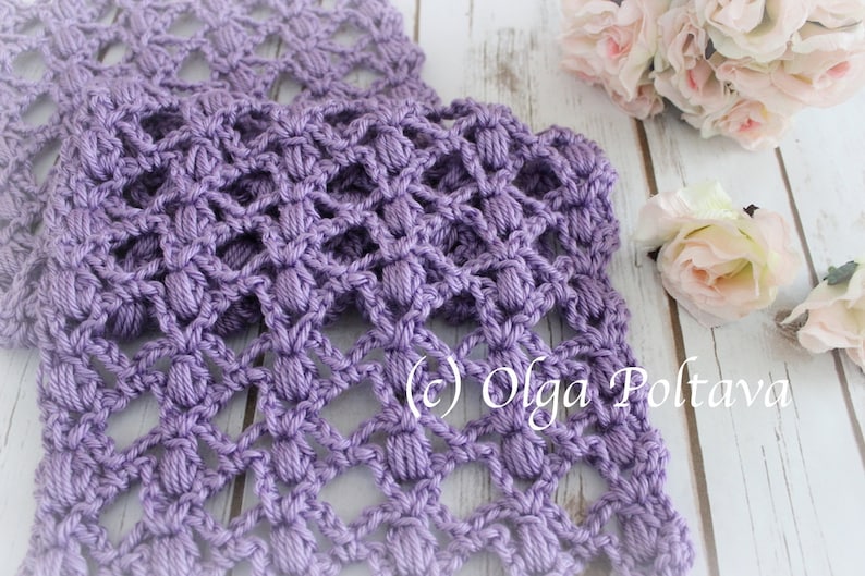 May include: A close-up of a handmade, lavender-coloured crochet scarf with a textured, openwork design. The scarf is made of soft yarn and features a repeating pattern of raised stitches. The background includes flowers and the text "(c) Olga Poltava".