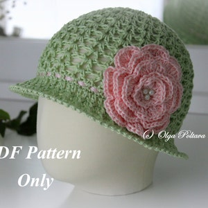 May include: A light green crochet hat with a pink flower and a small brim. The hat has a delicate open weave design and is adorned with a large, pink crocheted flower with pearl accents. The text "PDF Pattern Only" is visible.