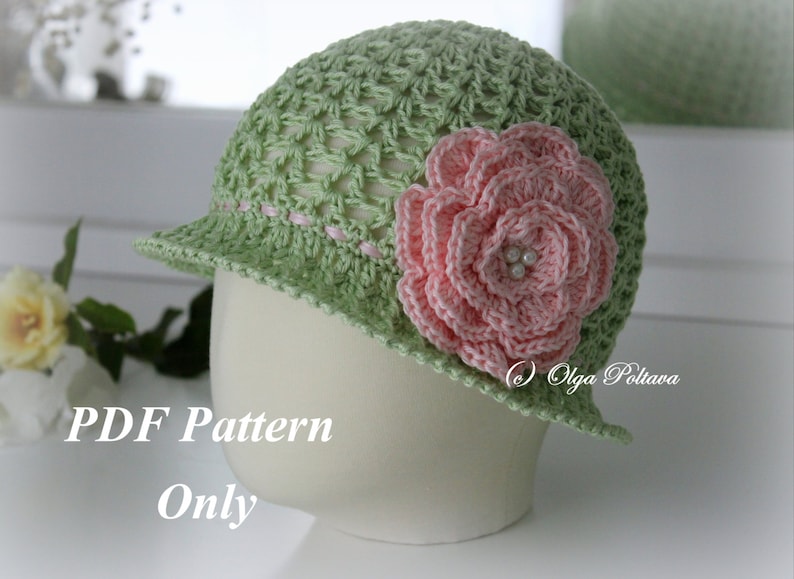 May include: A light green crochet hat with a pink flower and a small brim. The hat has a delicate open weave design and is adorned with a large, pink crocheted flower with pearl accents. The text "PDF Pattern Only" is visible.
