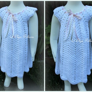 May include: White crochet dress with a pink ribbon bow at the neckline. The dress has a scalloped hem and is made with a delicate stitch pattern.