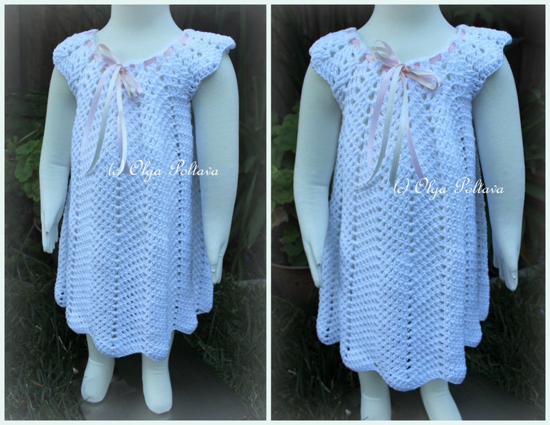May include: White crochet dress with a pink ribbon bow at the neckline. The dress has a scalloped hem and is made with a delicate stitch pattern.