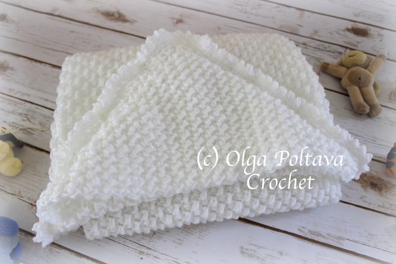 May include: White crocheted baby blanket with a ruffled edge. The blanket is folded in half and lying on a white wooden surface. (c) Olga Poltava Crochet