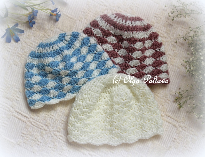 May include: Three handmade crochet baby hats in soft colours. One is light blue and white with a diamond pattern, another is white, and the third is burgundy and white. The hats are displayed on a white surface with floral accents.