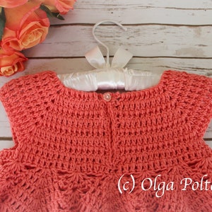 May include: Coral-colored crochet dress for a baby, displayed on a white hanger with a satin ribbon. The dress features a detailed stitch pattern and a small button closure. The background includes peach roses and a wooden surface.