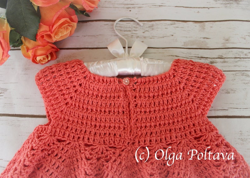 May include: Coral-colored crochet dress for a baby, displayed on a white hanger with a satin ribbon. The dress features a detailed stitch pattern and a small button closure. The background includes peach roses and a wooden surface.