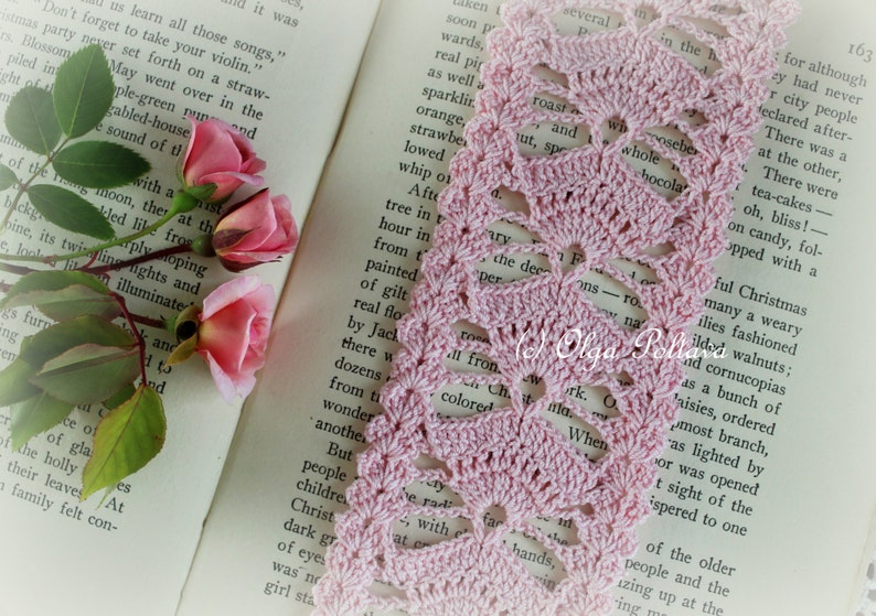 May include: A pink crocheted bookmark with a delicate floral pattern. The bookmark is laying on top of an open book with text.