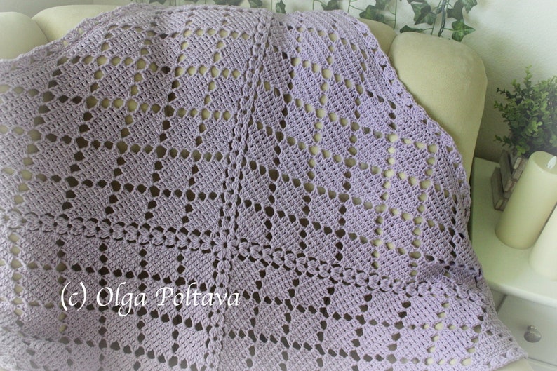 Puede incluir: Una manta de crochet de color lavanda con un delicado patr&oacute;n de calado. La manta est&aacute; colocada sobre un sof&aacute; beige claro.