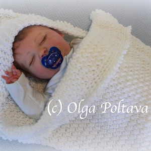 May include: A white crocheted baby blanket with a hood, featuring a textured stitch pattern. The blanket is wrapped around a sleeping baby doll with a pacifier.