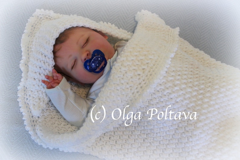 May include: A white crocheted baby blanket with a hood, featuring a textured stitch pattern. The blanket is wrapped around a sleeping baby doll with a pacifier.