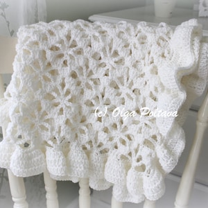 May include: A white crocheted baby blanket with a delicate floral pattern and a ruffled edge. The blanket is draped over a white chair.