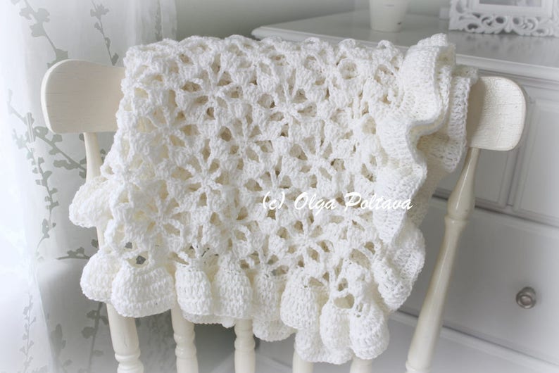 May include: A white crocheted baby blanket with a delicate floral pattern and a ruffled edge. The blanket is draped over a white chair.