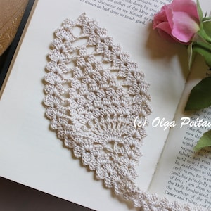 May include: A white crocheted bookmark with a delicate floral design, resting on an open book with a pink rose and green leaves. The bookmark is a beautiful handmade piece of art.