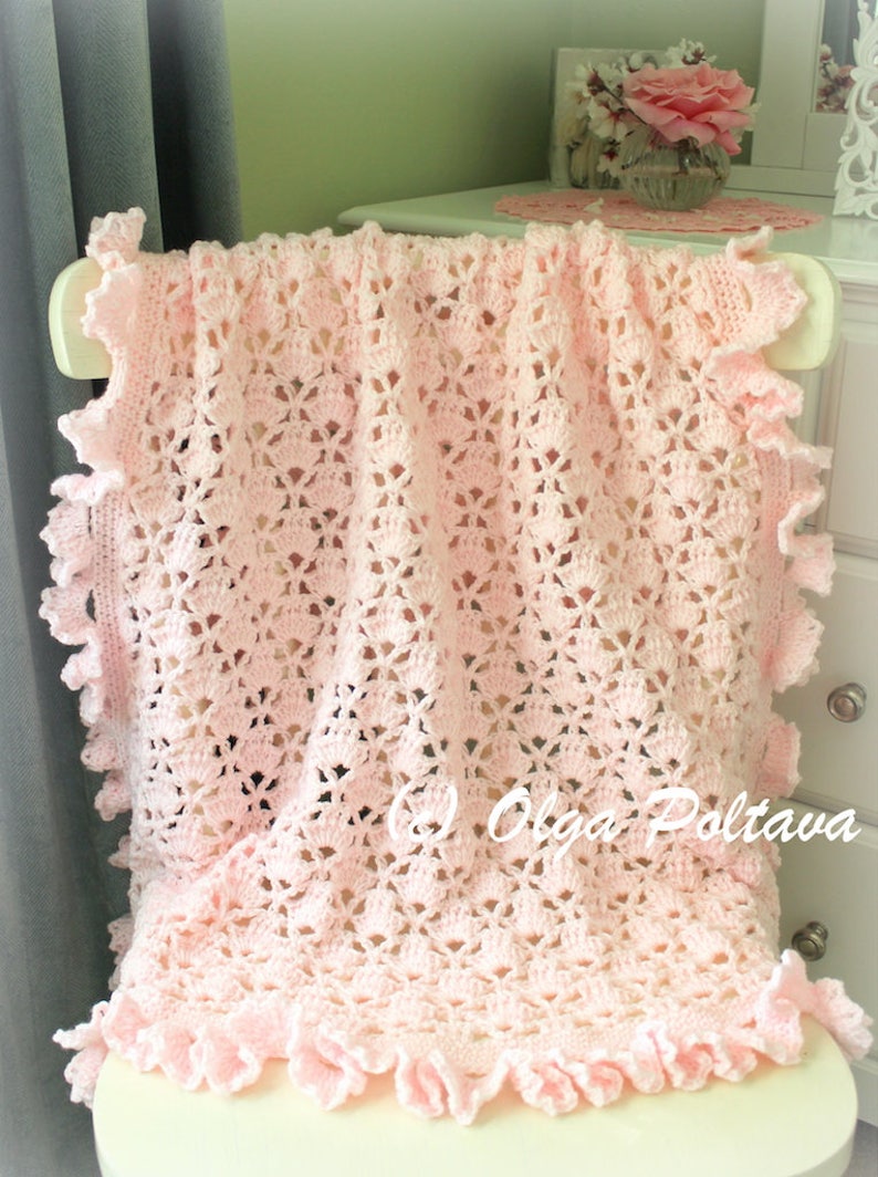 May include: A pink crocheted baby blanket with a delicate lace pattern and ruffled edges. The blanket is draped over a white chair.