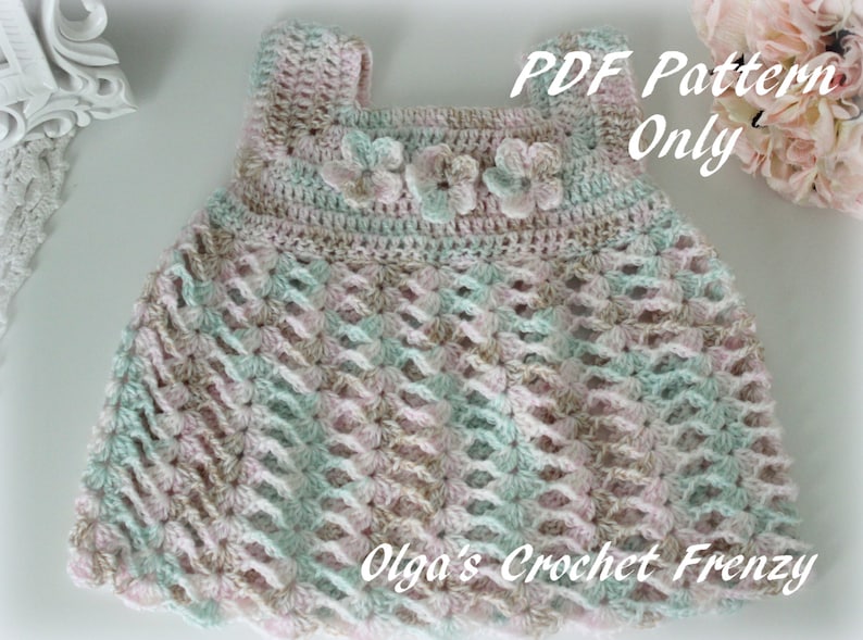 May include: A pastel-colored crochet baby dress with a scalloped hem. The dress features a square neckline and is adorned with three small flower appliqu&eacute;s. The text "PDF Pattern Only" is visible. "Olga's Crochet Frenzy" is also visible.