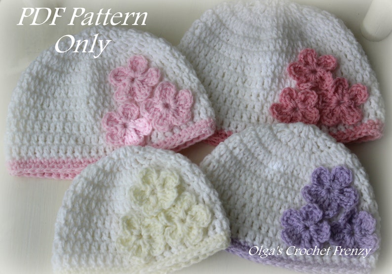 Op de afbeelding: Een verzameling witte gehaakte babyhoeden, elk versierd met een tros gehaakte bloemen in verschillende pastelkleuren. De hoeden hebben een roze of paarse rand. De tekst "PDF Pattern Only" is zichtbaar bovenaan.