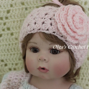 May include: A pink crocheted headband with a large pink flower on top. The headband is worn by a doll with brown hair and blue eyes.