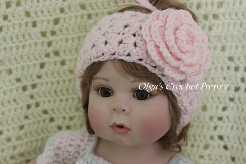 May include: A pink crocheted headband with a large pink flower on top. The headband is worn by a doll with brown hair and blue eyes.