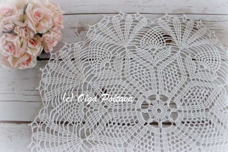 May include: White crocheted doily with a delicate, intricate design. The doily has a circular shape and features a central motif with radiating patterns.