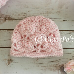 May include: A light pink crocheted hat with a floral pattern. The hat is handmade and features a scalloped edge. The text "(c) Olga Poltava" is visible on the hat. The hat is on a light wood surface.