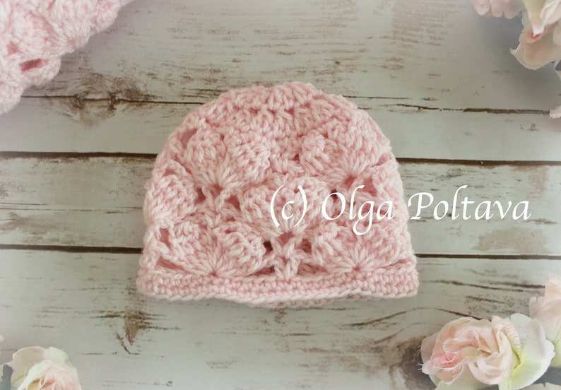 May include: A light pink crocheted hat with a floral pattern. The hat is handmade and features a scalloped edge. The text "(c) Olga Poltava" is visible on the hat. The hat is on a light wood surface.