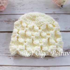 May include: A white crocheted baby hat with a textured, scalloped design. The hat is on a white wooden background. (c) Olga Poltava