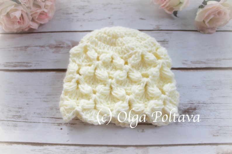 May include: A white crocheted baby hat with a textured, scalloped design. The hat is on a white wooden background. (c) Olga Poltava