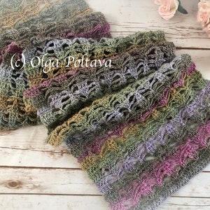 May include: A crocheted scarf in shades of green, purple, and brown with a lacy pattern. The scarf is folded in half and lying on a white wooden surface.