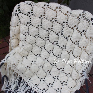 May include: A white crocheted throw blanket with a scalloped edge and fringe. The blanket is made with a textured stitch that creates a raised pattern. The blanket is draped over a black metal chair.