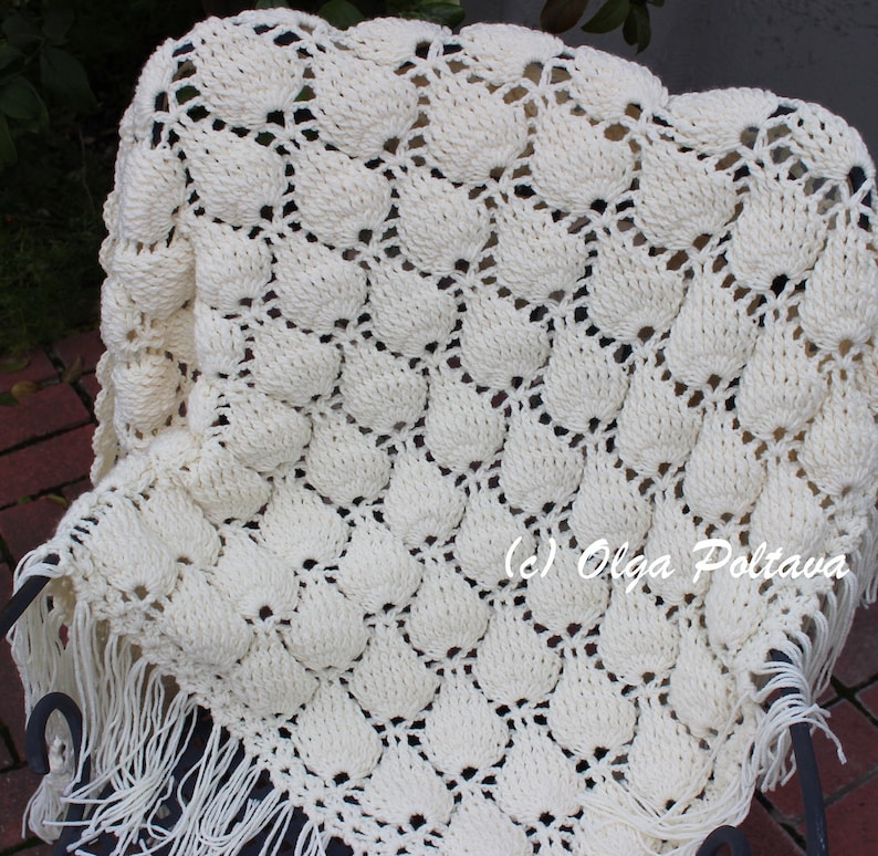 May include: A white crocheted throw blanket with a scalloped edge and fringe. The blanket is made with a textured stitch that creates a raised pattern. The blanket is draped over a black metal chair.