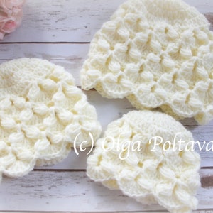 May include: Three crocheted hats in a cream color with a textured, raised pattern. The hats are arranged on a white wooden surface.