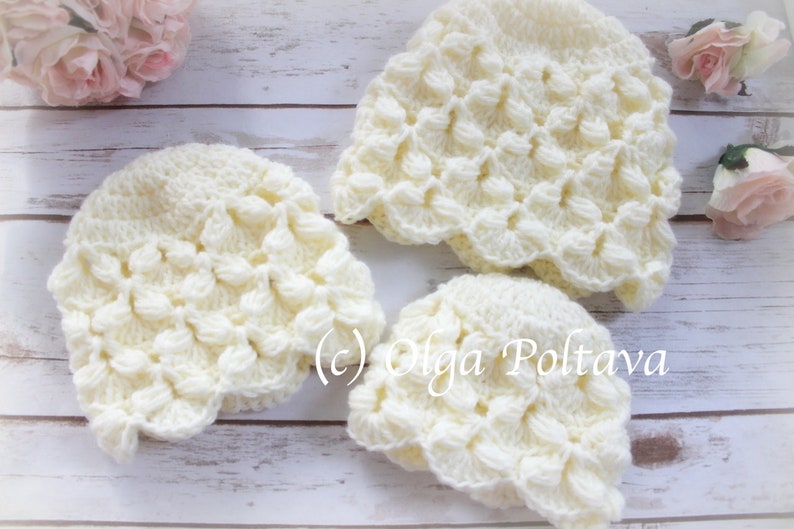 May include: Three crocheted hats in a cream color with a textured, raised pattern. The hats are arranged on a white wooden surface.
